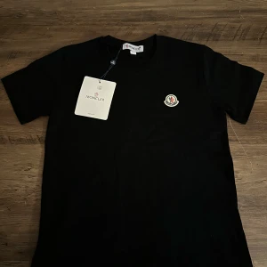 Svart Moncler t-shirt - Säljer en helt ny moncler tisha, på grund av att den ar for liten. Moncler storlek 150 men passar som xs/mindre xxs. Perfekt för dig som gillar stilrena och exklusiva plagg.