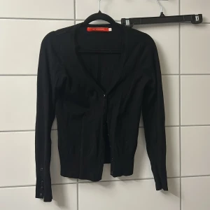 Svart kofta med knappar - En svart, klassisk kofta från H&M L.O.G.G. med lång ärm och knappar framtill. Snygg v-ringning och smal passform som sitter tajt mot kroppen. Perfekt att slänga över en t-shirt eller linne för en clean look.