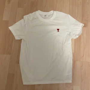 Vit t-shirt från AMI med rött hjärta - Vit t-shirt från AMI med klassisk passform och korta ärmar. På bröstet finns ett litet rött hjärta med bokstaven A broderat. Tillverkad i mjuk bomull och perfekt för en clean och stilren look.
