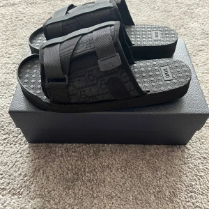 Svarta dior alpha slides  - Ett par stilrena svarta Dior alpha sandaler. Perfekt för varma dagar eller som ett elegant tillskott till din garderob. Sandalerna har en distinkt design med justerbara remmar och Dior-logotypen.