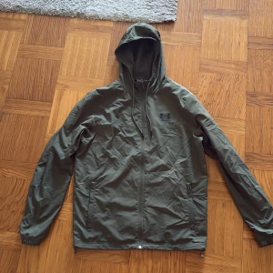 Olivgrön vindjacka från Under Armour - Säljer typ en helt ny olivgrön vindjacka från Under Armour med huva och dragkedja framtill. Jackan har meshfoder, två sidofickor med dragkedja och elastiska muddar vid ärmsluten. Perfekt för blåsiga dagar och sportiga outfits. Använder inte längre. Vill få bort så fort som möjligt!!!
