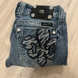 Miss Me jeans med strass och brodyr - Snygga blå Miss Me jeans med slitningar och stora broderade detaljer med strass på bakfickorna. Jeansen har raka ben, silverfärgade knappar och coola detaljer som ger en unik look. Perfekta för dig som vill sticka ut med din stil.
