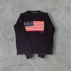 Svart stickad tröja Polo Ralph Lauren - Svart stickad tröja från Polo Ralph Lauren med amerikansk flagga och RL-broderi på bröstet. Klassisk rund halsringning och ribbade muddar vid ärmslut och nederkant. Perfekt för dig som gillar preppy stil. Säljer den då den  ej passar mig/ lite liten i storleken. Pris går att diskutera. Ralph lauren posen följs med och taggen. 