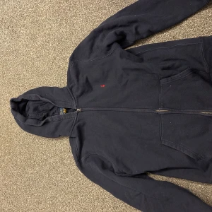 Svart hoodie från Polo Ralph Lauren  - Mörkblå hoodie från Polo Ralph Lauren.Klassisk design med dragkedja, huva och kängurufickor. Broderad röd logga på bröstet. Mjuk insida i bomullsmix, perfekt för höst och vinter. Enkel och stilren, passar till allt. Storlek XL i barn så passar S men även XS