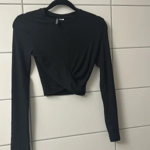 Svart omlottopp med ribbad struktur - Snygg svart långärmad topp  med ribbad struktur och omlottdetalj framtill. Croppad modell som ger en trendig look, perfekt att matcha med höga jeans eller kjol. Enkel rund halsringning och smal passform.