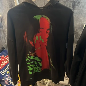 Svart Billie Eilish hoodie med tryck - Svart hoodie med stor färgstark Billie Eilish-porträtt på framsidan i rött och grönt samt rött Billie Eilish-tryck på ryggen. Hoodien har huva och långa ärmar. Den är u skönt material och är jätte fin