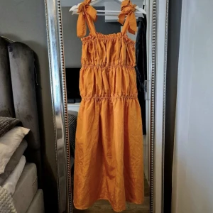 Orange sommarklänning med rosettband - Superfin orange långklänning med smockad överdel och breda axelband som knyts i stora rosetter. Klänningen har volangig kjol och är perfekt för varma dagar. Lätt och luftig känsla, passar dig som gillar färg och en avslappnad stil.