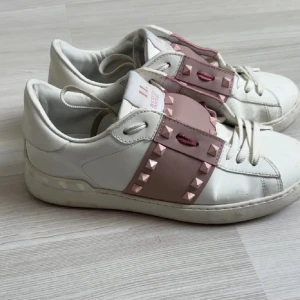 Valentino Rockstud sneakers i vitt och rosa - Snygga Valentino Rockstud sneakers i vitt skinn med breda ljusrosa band och ikoniska nitar över vristen. Klassisk rund tå, vita snören och platt sula. Perfekta för dig som vill sticka ut med en lyxig och edgy vibe.