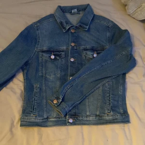 Klassisk blå jeansjacka - Snygg blå jeansjacka med klassisk passform och silverfärgade knappar framtill. Jackan har två bröstfickor med lock och knappar samt två sidofickor. Perfekt att slänga över en hoodie eller t-shirt för en chill look.