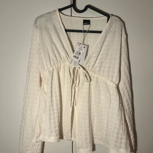 Vit blus med knyt från Gina Tricot - Supersöt vit blus från Gina Tricot med v-ringning och knyt framtill. Lätt strukturerat tyg och långärmad modell som ger en luftig och chill vibe. Perfekt att styla med jeans eller kjol för en fräsch look.