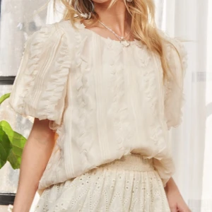 Romantisk volangblus i ljus beige - Supersöt blus i ljus beige med puffiga korta ärmar och volangdetaljer längs hela toppen. Blusen har rund halsringning och ett skirt, luftigt tyg som ger en romantisk vibe. Perfekt att styla med kjol eller jeans för en trendig look.