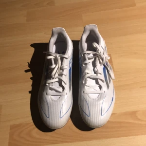 Vita Adidas F50 strl 41  - Säljer ett par vita Adidas fotbollsskor med blå och grå detaljer. Skorna har snörning och mönstrad sula med dobbar för bra grepp på planen. Tillverkade i syntetmaterial och designade för att vara lätta och snabba. Perfekta för dig som vill sticka ut på fotbollsplanen.