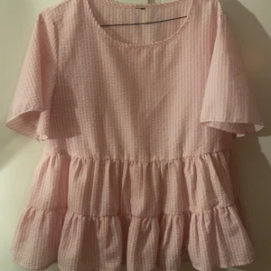 Rosa volangblus med kort ärm - Supersöt rosa blus med  randigt mönster och flera volanglager nedtill. Kommer ej till användning. Funkar för inprincip alla storlekar, men ungefär S - L. Materialet känns lätt och luftigt, vilket gör den skön att bära under varmare dagar!🥰 aldrig använd.