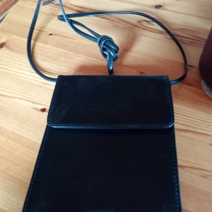 Svart axelväska i skinn från Lisa and Co - En stilren svart axelväska från Lisa and Co i slätt skinn. Väskan har ett minimalistiskt uttryck med lock och lång axelrem. Perfekt storlek för mobil och småprylar, och enkel att matcha till olika outfits. Måtten 20 cm hög och 16 cm bred.