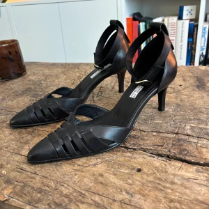 Nya Blankens Klackar The Amanda - Blankens i modell: The Amanda! Nya oanvända stilrena svarta pumps i skinn med spetsig tå, flätade detaljer framtill och smal ankelrem med spänne. Snygg klack som ger en elegant siluett. Perfekta för dig som gillar klassisk men modern stil. Nypris: 2700kr