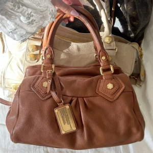 Chestnut Q Classic Baby Groove! - Så snygg klassisk chestnut färgad handväska i nyskick från Marc by Marc Jacobs i äkta läder med gulddetaljer och dubbla handtag. Väldigt snygg färg som passar till allt, den perfekta sommarväskan som rymmer mycket men är smidig och nätt. Lilla modellen Q Classic. Inga repor eller hål på väskans läder förutom ett litet märke, syns på bilderna. Otroligt välbevarad vintage modell! Perfekt för att ge en stilren touch till din outfit. Skriv privat för fler bilder. Mått 34x18x12 (undersida)