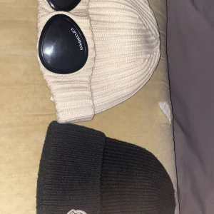 Gogglesmössa G.P. Company & svart Moncler - Kan skicka mer bilder pv, pris kan diskuteras.Två ribbstickade mössor: en beige G.P. Company gogglesmössa med svarta glasögon på uppviket och en svart klassisk mössa med broderad logga framtill. Båda är perfekta för kalla dagar och har en modern streetwear-vibe.