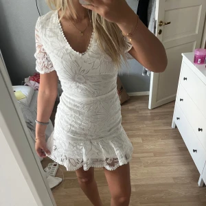 Vit kort spetsklänning med öppen rygg - Superfin vit kort klänning med blommigt spetsmönster, v-ringning och volangkant nertill. Klänningen har korta ärmar i transparent spets och en snygg öppen rygg med korsade band. Perfekt för sommarens alla tillfällen!