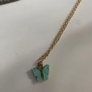 Guldhalsband med turkos fjärilshänge - Snyggt halsband med guldfärgad kedja och ett hänge i form av en turkos fjäril. Fjärilen har en mjuk, pastellig färg och ger ett lekfullt intryck. Perfekt accessoar för att lyfta din outfit med en söt detalj.
