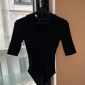 Svart bodysuit med krage från Zara - Svart bodysuit från Zara med klassisk krage och knappar framtill. Modellen har trekvartsärm och är figurnära med en stilren look. Perfekt att styla med jeans eller kjol för en clean vibe. Helt ny