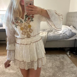 Vit broderad kortklänning med volanger - Superfin vit kortklänning med broderade blommor och mönster i gult och rosa. Klänningen har v-ringning, volangärmar och flera lager volanger i kjolen. Materialet är bomull med spetsdetaljer och broderier. Perfekt för sommarens alla tillfällen!