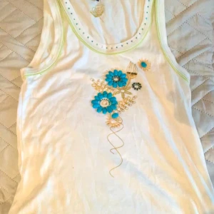 Vit linnetopp med blå blommor CN Collection - Supersöt vit linnetopp från CN Collection med unika blå blommor och guldbroderi framtill. Toppen har ärmlös design och små blå stenar runt ringningen för extra detalj. Perfekt för dig som gillar färgglada och kreativa plagg inför sommaren 😛