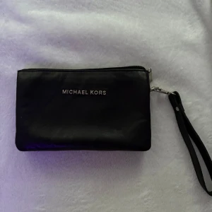 Svart Michael Kors kuvertväska i skinn - Snygg svart kuvertväska från Michael Kors i äkta skinn med silverfärgad dragkedja och avtagbar handledsrem. Insidan har flera fack och ett innerfoder med MK-logga. Perfekt för att bära det viktigaste och ge en clean look till din outfit. Pris kan diskuteras.