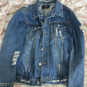 Blå jeansjacka med slitningar  - Säljer en klassisk blå jeansjacka från bikbok med coola slitningar på ärmarna och vid nederkanten. Jackan har knappar framtill, två bröstfickor med lock och en snygg krage. Perfekt för att lyfta din outfit! Passar även en S lite oversize jacka. GE PRISFÖRSLAG!💕