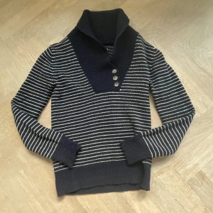 Randig blå ulltröja från Sacoor Brothers - Mörkblå och vitrandig stickad tröja från Sacoor Brothers i 100% lammull. Tröjan har hög krage med tre knappar framtill, breda ribbade muddar och en klassisk, lite loose passform. Perfekt för kalla dagar när du vill vara både snygg och bekväm. Tveka inte på att höra av dig!!!