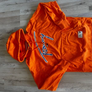 Orange hoodie från Tommy Jeans - Snygg orange hoodie från Tommy Jeans med stor blå och vit logga framtill. Tröjan har huva med dragsko, magficka och ribbade muddar. Perfekt för dig som vill sticka ut med färg och streetstyle. Materialet är mjuk bomull som känns skönt mot huden.