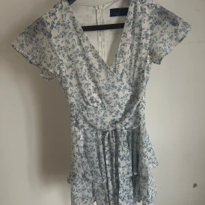 Blommig vit och blå kortklänning - Supersöt kortklänning i vitt med blått blommönster. Klänningen har v-ringning, volangdetaljer och knyt i midjan, shorts är inbyggt. Lätt och flowy känsla med korta ärmar, perfekt för varma dagar. Strl XXS/32.