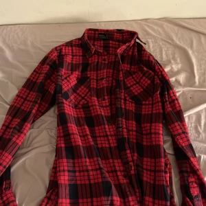 Röd rutig skjorta från Urban Classics - Säljer en röd och svart rutig skjorta från Urban Classics i storlek S. Skjortan är tillverkad i 100% bomull, har långa ärmar, klassisk krage och knäppning framtill. Perfekt för dig som gillar en avslappnad och tidlös stil.
