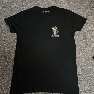 Svart Amiri t-shirt med ängeltryck - Svart t-shirt från Amiri med stort tryck av en ängel och gröna blad på ryggen samt mindre tryck på bröstet. Klassisk passform med rund hals och korta ärmar. Snygg och stilren design som sticker ut med detaljerad grafik.