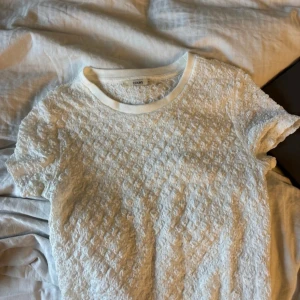 Vit spetsmönstrad t-shirt från Colin's - Supersöt vit t-shirt från Colin's med korta ärmar och ett unikt spetsliknande mönster över hela tyget. Rund halsringning med ribbad kant. Perfekt för dig som gillar en romantisk och fräsch stil.