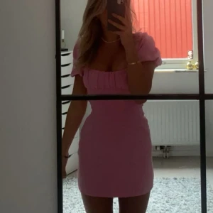 Rosa kort puffärmad klänning - Supersöt rosa kortklänning med puffärmar och fyrkantig urringning. Klänningen har öppen rygg med snörning som ger en snygg detalj. Figurnära passform och perfekt för dig som vill sticka ut lite extra.