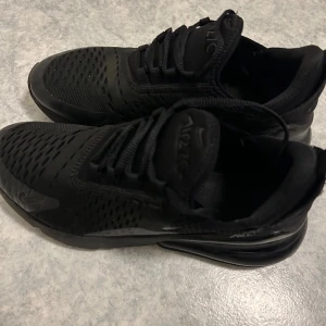 Svarta Nike Air Max 270 sneakers - Säljer ett par helsvarta Nike Air Max 270 sneakers med snygg mesh-överdel och klassisk Air Max-sula. Skorna har snörning och en sportig vibe, perfekt för dig som gillar streetstyle. Luftig ovandel i syntet och textil för extra komfort.