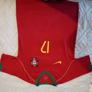 Portugal C. Ronaldo 17 Nike fotbollströja - Säljer en klassisk röd Portugal fotbollströja från Nike med nummer 17 och C. Ronaldo på ryggen. Tröjan har gula detaljer, gröna muddar och Portugals landslagsemblem på bröstet. Perfekt för dig som älskar fotboll och Ronaldo.