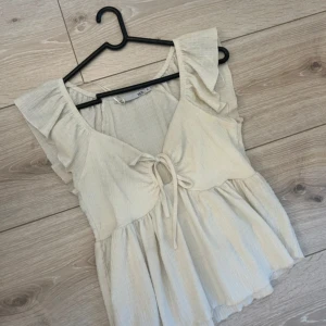 Beige blus med volang och knyt - Supersöt beige blus med volangdetaljer på axlarna och knyt framtill. Blusen har v-ringning och är tillverkad i ett lätt, luftigt bomullstyg. Perfekt för varma dagar och ger en romantisk vibe till din outfit. 