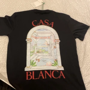 Svart t-shirt från Casablanca - Svart t-shirt från Casablanca med färgglad tryckt illustration av en portal och landskap på både bröstet och ryggen. Röd text 'CASA BLANCA' ger en snygg kontrast. Klassisk passform och rund hals, perfekt för dig som gillar stilrena men unika plagg.