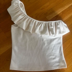 Vit oneshoulder topp med volang - Såå fin och populär volang topp från H&M! Toppen är knappt använd, i väldigt fint skick förutom någon typ av fläk (syns knappt), inte tvättat den men skulle tro att den går bort i en tvätt (se sista bilden)❤️❤️ Man kan ha den åt två olika hål beroende på vilket håll man vill ha volangen på!🌟