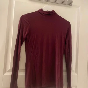 Vinröd polotröja i tunn bomull - Vinröd polotröja med lång ärm och tight passform. Tunt och mjukt bomullsmaterial som känns skönt mot huden. Perfekt att styla lager på lager eller bära som den är. Enkel och stilren design utan mönster eller detaljer.