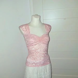 Rosa spetsig blus med breda axelband - Supersöt ljusrosa blus i spets med breda axelband och lätt rynkad byst. Toppen är figurnära och har ett romantiskt blommönster i spetsen. Perfekt för dig som gillar en feminin och trendig look.