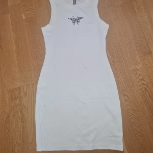 Vit ärmlös ribbad kortklänning - Säljer en vit, tight och ärmlös ribbad kortklänning med tribaltryck framtill vid halsen. Klänningen har en sportig och enkel look, perfekt för dig som gillar minimalistisk stil med en edgy detalj.