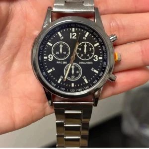 Herrklocka - Herrklocka med svart urtavla, silverfärgat armband i metall och kronograf-funktion. Klockan har tydliga vita markeringar, tre mindre urtavlor och en gul detalj på kronan. Fick i present men är för stor, helt ny.