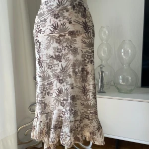 Beige blommig långkjol med volang - Supersöt långkjol i beige och bruna toner med blommigt mönster. Kjolen har en figurnära passform och avslutas med en volangkant nertill som ger en romantisk vibe. Tunt, lätt material som känns perfekt för vår och sommar.