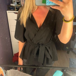 Svart blus med knytband - Snygg svart blus med v-ringning och breda, korta ärmar. Blusen har ett knytband i midjan som ger en markerad siluett och ett flowigt fall nedtill. Perfekt att styla med jeans eller kjol för en trendig look.
