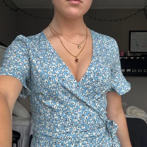 Blå blommig omlottklänning - Supersöt blå kort omlottklänning med vitt blommönster och volangkant. Klänningen har v-ringning, knyt i midjan och korta ärmar med lätt volang. Perfekt för sommarens alla tillfällen och ger en fräsch och ungdomlig vibe.