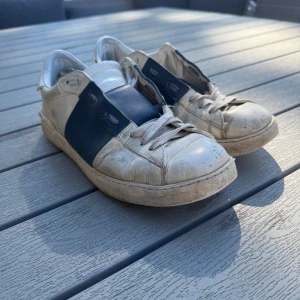 Valentino Open sneakers vit/blå skinn - Snygga Valentino Open sneakers i vitt skinn med bred blå rem över mitten. Klassisk rund tå, vita snören och platt sula. Perfekta för dig som gillar stilrena och exklusiva sneakers med en twist. Diskret logga på hälen och insidan.