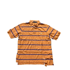 Ralph Lauren piké  - Orange pikétröja från Polo Ralph Lauren i storlek XLT. Tröjan har blå horisontella ränder, klassisk krage och två knappar framtill. Broderad logga på bröstet. Tillverkad i bomull och har korta ärmar. Perfekt för en avslappnad och stilren look. Se defekt på sista bilden ! Jag skickar ej bild när jag bär plaggen och jag vet ej vilken längd och vikt de passar då jag inte prövar kläderna jag säljer 😃