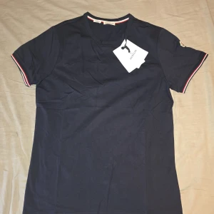 Moncler t-shirt slim fit - Snygg t-shirt från Moncler i slim fit-modell. Klassisk rund halsringning, korta ärmar med röd, vit och blå rand på mudden samt Moncler-logga på ärmen. Tillverkad i mjuk bomull för en skön känsla. --- M/L (slim fit) 